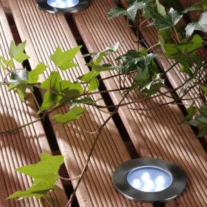 Brevus Decklight with stainless steel finish installed on a wooden deck surrounded by ivy, showcasing warm white LED lighting.